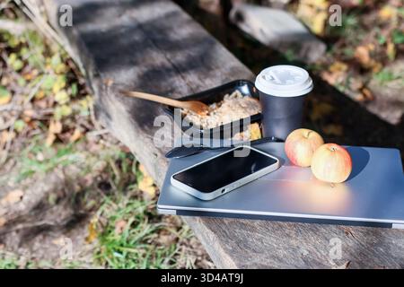 Nahaufnahme der Getreideschale im Behälter, Kaffee, Äpfel, Utensilien, Telefon auf Laptop draußen. Draufsicht des offenen schwarzen Behälters mit Getreidefutter Stockfoto