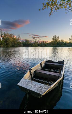 Ein einzelnes kleines Boot schwimmt auf einem ruhigen See unter einem pastellfarbenen Sonnenuntergang Himmel umgeben von Bäumen. Stockfoto