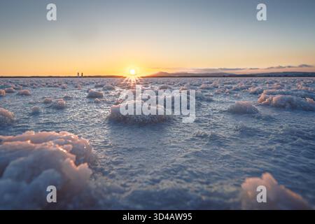 Salzkristalle auf der Oberfläche eines Salzsees bei Sonnenuntergang, die Sonne scheint tief am Horizont. Stockfoto