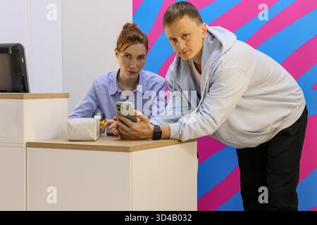 Mann und Frau untersuchen das Smartphone am Service-Schalter. Zwei Personen lehnen sich über den Tresen, um den Bildschirm des Telefons in farbenfroher Inneneinrichtung zu sehen. Stockfoto