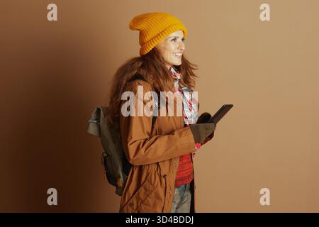 Eine junge Frau in gelber Mütze und brauner Jacke lächelt im Profil, während sie ein Tablet benutzt und zeigt den modernen Reisenden oder Pendler, der mit ihr verbunden ist Stockfoto