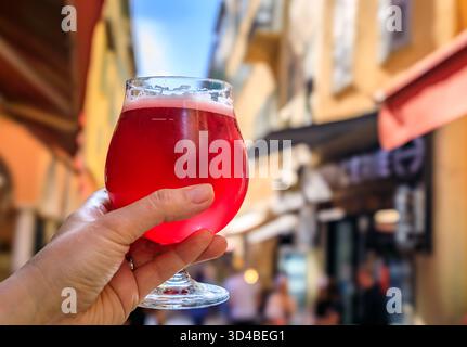 Hand hält ein Glas Himbeer lambic Ale im Freien mit einem Hintergrund verschwommener Gebäude in der Altstadt oder Vieille Ville in Nizza, Südfrankreich Stockfoto