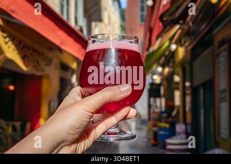 Hand hält ein Glas Himbeer lambic Ale im Freien mit einem Hintergrund verschwommener Gebäude in der Altstadt oder Vieille Ville in Nizza, Südfrankreich Stockfoto