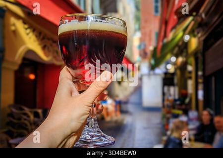 Hand hält ein Glas Bier im Freien mit verschwommenen Gebäuden in der Altstadt oder Vieille Ville in Nizza, Südfrankreich Stockfoto