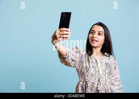 Eine lächelnde, selbstbewusste Person macht Fotos mit ihrer Handy-App, fühlt sich hübsch an und macht Fotos vor blauem Hintergrund. Eine junge indianerin zeigt Freude und eine positive Einstellung. Stockfoto