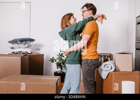 Glückliche Paarumarmungen umgeben von Umzugskästen, während des Umzugs, einen freudigen Moment miteinander teilen, in ein neues Haus ziehen Stockfoto