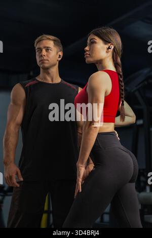 Paare stehen stolz in einem modernen Fitnessstudio, das ihre Fitness und ihren Stil zeigt. Der Mann trägt ein schwarzes ärmelloses Hemd, während die Frau ein rotes Crop-Top mit schwarzen Leggings trägt. Stockfoto