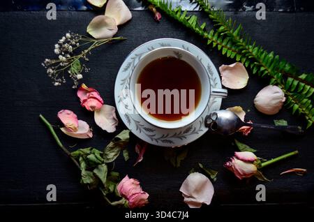 Ein stimmungsvolles, dunkles, florales Stillleben mit einer Porzellanteasse gefüllt mit reichhaltigem, dunklem Tee, umgeben von zarten rosa und roten Rosenblüten, getrockneter Fluss Stockfoto