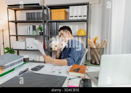 Junger männlicher Architekt, der durchdacht am Schreibtisch arbeitet, Hauspläne und Baupläne analysiert und Details sorgfältig überprüft, um sicherzustellen, dass Stockfoto