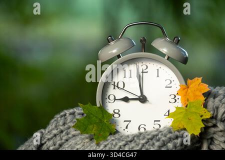 Eine Nahaufnahme eines klassischen Weckers, umgeben von bunten Herbstblättern, auf einem weichen, gestrickten Schal mit verschwommenem grünem Hintergrund. Stockfoto