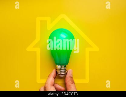 Hand hält grüne Glühbirne mit Haussymbol auf gelbem Hintergrund, symbolisiert Energiesparen, umweltfreundliches Zuhause, grüne Elektrizität, nachhaltiges Leben Stockfoto