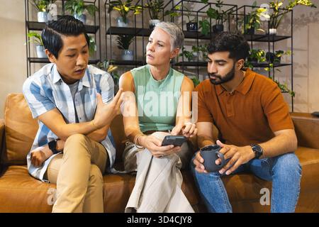 Über die Arbeit diskutieren, verschiedene Kollegen sitzen auf der Couch in der Bürolounge mit Smartphone und Kaffeetasse Stockfoto
