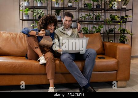 Stöbern Sie durch verschiedene Paare, die einen Laptop auf brauner Ledercouch im Wohnzimmer mit Kaffeetassen bewerten Stockfoto
