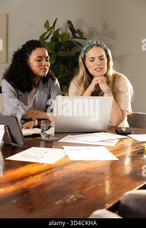 Verschiedene weibliche Kollegen, die Karten auf silbernem Laptop im Büro mit Notebook und Stift überprüfen Stockfoto