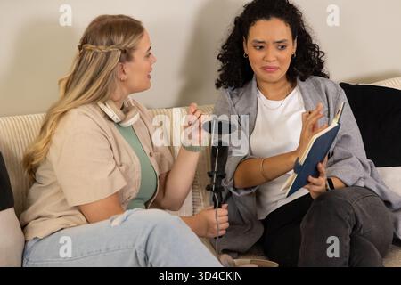 Verschiedene weibliche Co-Moderatoren, die Podcasts auf beigefarbenem Sofa im Heimstudio aufnehmen, mit Mikrofon-Pop-Filter Stockfoto