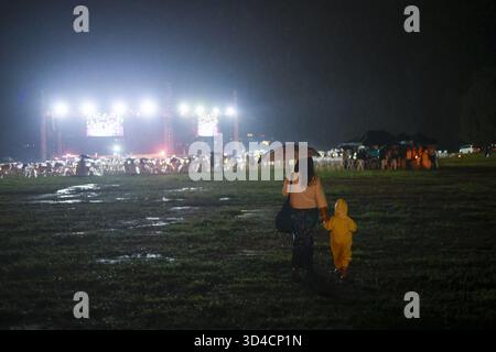 Liebevolle Mutter und Kind gehen bei einem Outdoor-Musikfestival bei Nacht bei Regen. Das Kind trägt einen gelben Regenmantel, während die Frau einen Regenschirm hält, der Schutz bietet Stockfoto