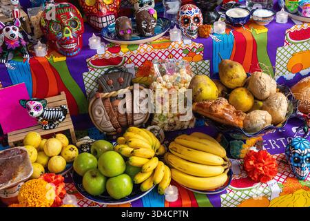 Ein Tisch ist mit einer farbenfrohen Tischdecke und einer Vielzahl von Obst und Gemüse, einschließlich Bana, bedeckt. Mexikanischer Tag der Toten Tradition, Opfergaben, fo Stockfoto