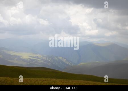 Weite Aussicht auf riesige, mehrschichtige, blau-graue Bergkämme und sanfte grüne Hügel unter einem dramatischen, stimmungsvollen, bewölkten Himmel mit atmosphärischem Licht Stockfoto