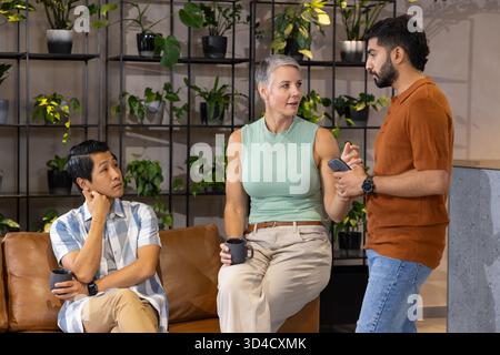Sprechen über Kaffeetassen, verschiedene Kollegen sitzen auf dem Sofa in der Bürolounge, mit Smartphone Stockfoto