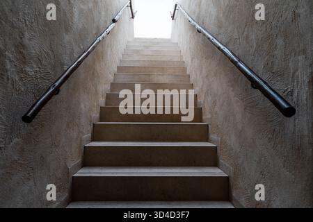 Ein dramatischer Blick nach oben auf eine moderne Treppe, die aus der Dunkelheit in einen hellen, gut beleuchteten Raum übergeht. Das Bild erfasst den Kontrast zwischen schattigen Stufen und dem Licht oben an der Treppe Stockfoto