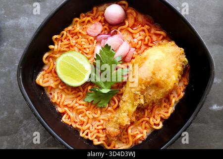 Blick von oben auf scharfe Hähnchenramen-Nudeln mit knusprig gebratenem Drumstick, Limette und Schalotten in einer schwarzen Schüssel. Ein herzhaftes und wohltuendes asiatisches Gericht, reich an f Stockfoto