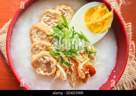 Dies ist eine Nahaufnahme von traditionellem indonesischem Frühstück oder Komfort-Essen, bekannt als Bubur Ayam, serviert in einer roten Schüssel mit verschiedenen köstlichen, herzhaften Toppi Stockfoto