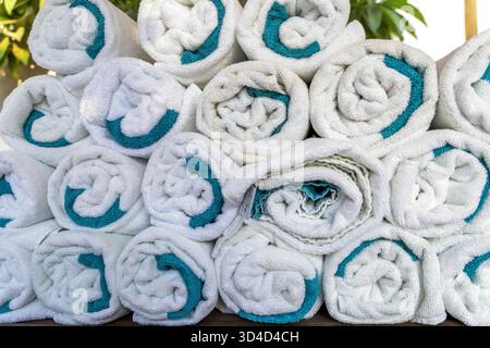Nahaufnahme von säuberlich gerollten weißen Handtüchern mit türkisfarbenen Kanten, pyramidenförmig gestapelt. Perfekt für Spa-, Hotel- oder Resort-Themen, die Sauberkeit, Komfort und Entspannung symbolisieren Stockfoto