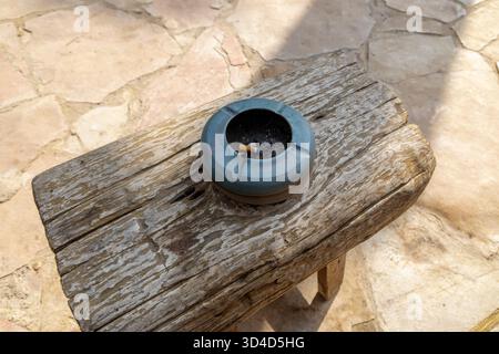 Benutzte Zigarette in einem blauen Aschenbecher auf einer verwitterten Holzbank. Die Bank steht auf einer sonnendurchfluteten Steinterrasse und schafft eine rustikale Atmosphäre im Freien mit natürlichen Texturen und Schatten Stockfoto