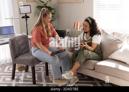 Vielfältige Freundinnen unterhalten sich im gemütlichen Wohnzimmer mit Laptop, Ringlicht und Pflanzen Stockfoto