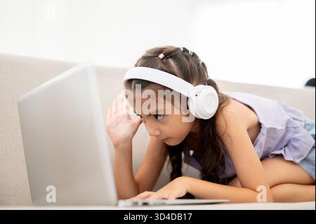 Ein asiatisches Kind oder ein Mädchen mit Kopfhörern schaut auf ihren Laptop und hebt eine Hand, während er sich auf einem Stoffsofa hockt. Video oder Videoanruf ansehen. Stockfoto