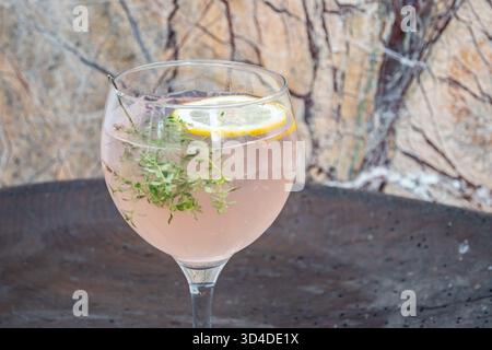 Hellrosafarbenes alkoholisches Sommergetränk in einem Weinglas mit Eis, Zitronenscheibe und frischem Thymian auf einem Heimtisch. Stockfoto