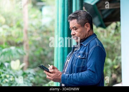 Ein älterer asiatischer Mann steht draußen in Indonesien und benutzt ein Smartphone, während er eine Zigarette hält. Ein offenes Lifestyle-Bild, das Technologie und Gewohnheit kombiniert. Stockfoto