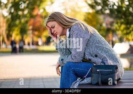 Porträt einer erschöpften Geschäftsfrau im Mantel, die Kopfschmerzen hat, während sie allein auf der Bank im Stadtpark sitzt. Stockfoto