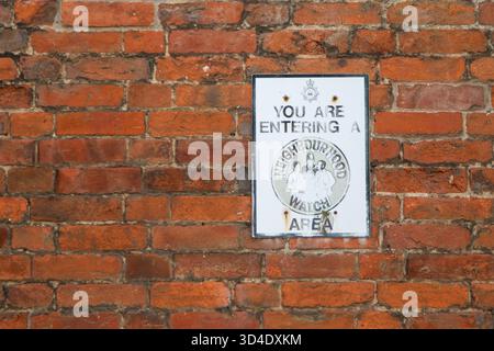 Auf einer Mauer aus weichen roten Suffolk-Ziegeln ist ein Warnschild aus der Nachbarschaft so verblasst und ausgewaschen, dass man befürchtet, dass es keine Abschreckung mehr ist, wenn es jemals so war Stockfoto