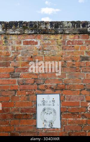Auf einer Mauer aus weichen roten Suffolk-Ziegeln ist ein Warnschild aus der Nachbarschaft so verblasst und ausgewaschen, dass man befürchtet, dass es keine Abschreckung mehr ist, wenn es jemals so war Stockfoto