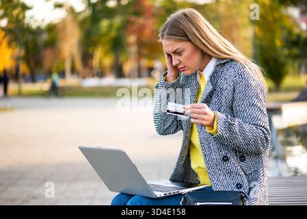 Geschäftsfrau im Mantel, die gestresst aussah, während sie einen Laptop benutzte und eine Kreditkarte hielt. Sie hat mit finanziellen Problemen zu kämpfen, während sie im Park arbeitet. Stockfoto