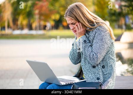 : Porträt einer überlasteten und gestressten Geschäftsfrau mit Kopfschmerzen, die mit dem Laptop auf einer Parkbank in der Stadt sitzt. Stockfoto