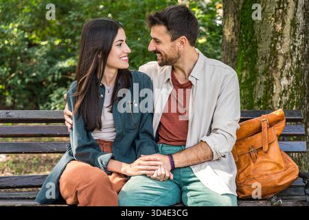 Junges Paar kuschelt auf einer Parkbank, lächelt und lehnt sich zu einem Kuss, teilt eine liebevolle Umarmung in sonnigen grünen Natur-Freizeitoutfits, warm C Stockfoto