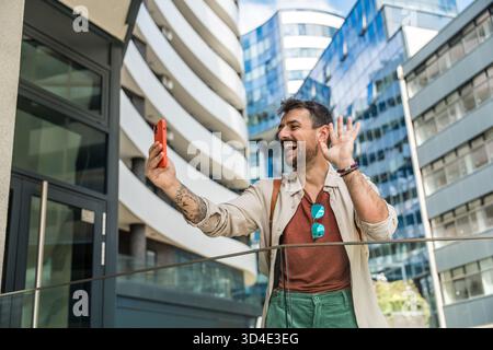 Junger Reisender lacht während eines Selfies in einem modernen Geschäftsviertel fröhlicher Millennial-Mann mit Rucksack, der urbane Architektur und Zitat einfängt Stockfoto