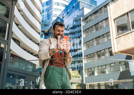 Junger Reisender lacht während eines Selfies in einem modernen Geschäftsviertel fröhlicher Millennial-Mann mit Rucksack, der urbane Architektur und Zitat einfängt Stockfoto