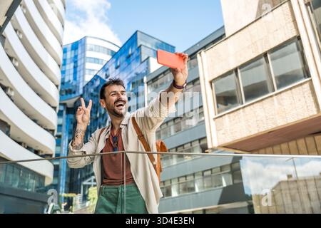 Junger Reisender lacht während eines Selfies in einem modernen Geschäftsviertel fröhlicher Millennial-Mann mit Rucksack, der urbane Architektur und Zitat einfängt Stockfoto