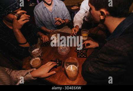 Freunde versammeln sich an einem Holztisch und spielen Karten mit Getränken während eines Abends Stockfoto