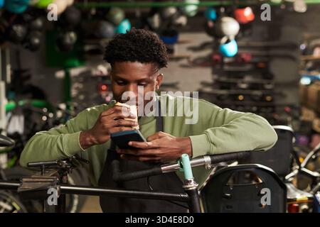 Junger Schwarzer isst Sandwich, während er das Smartphone im Workshop benutzt Stockfoto