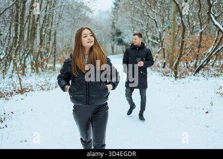 Ein charmantes Paar geht Hand in Hand durch einen verschneiten Wald, wo die elegant gekleidete Frau und ihr verspielter Partner Freude und Lachen in Winte teilen Stockfoto