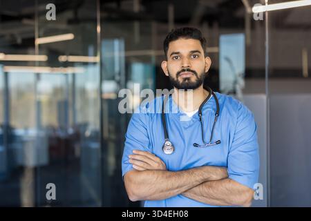 Indischer männlicher Arzt in blauen Peelings und Stethoskop, der selbstbewusst mit gekreuzten Armen steht und Professionalität und Hingabe in einer modernen Gesundheit darstellt Stockfoto