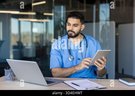 Junger männlicher Arzt, der blaue Peelings und Stethoskop trägt, an einem Schreibtisch sitzt und aktiv ein digitales Tablet für Gesundheitsdaten verwendet, in einer sauberen, verachtenden Weise Stockfoto
