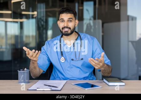 Männlicher Arzt in blauen Peelings und Stethoskop arbeitet an einem Schreibtisch, Blick auf die Kamera und Gesten während der Bereitstellung von Telemedizin-Service oder Gesundheitswesen A Stockfoto