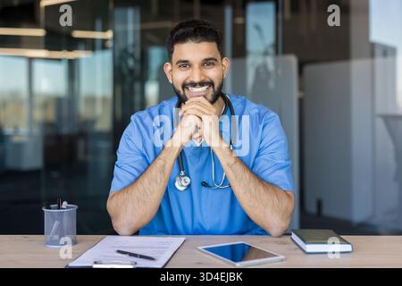 Junge indische Ärztin mit Peelings und einem Stethoskop, sitzt an einem Schreibtisch und lächelt selbstbewusst, verkörpert professionelle medizinische Versorgung und Unterstützung Stockfoto