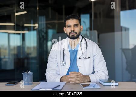 Selbstbewusster indischer Arzt in weißem Mantel mit Stethoskop, sitzt an einem modernen Klinikpult, blickt ruhig in die Kamera, vermittelt Professionalität, Exp Stockfoto