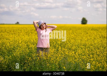 Frau mit Eimerhut steht auf gelbem Rapsfeld und genießt die Wärme und Ruhe eines sonnigen Sommertages Stockfoto